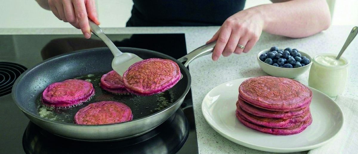 Vivid pink beetroot pancakes cooking in a frying pan, with a stacked plate, blueberries and yoghurt nearby.